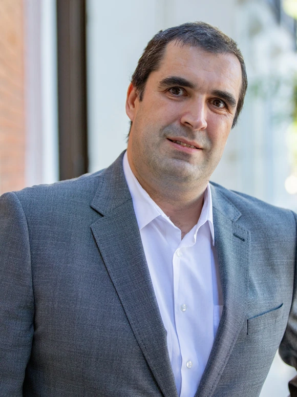 The image shows a smiling middle-aged man in a gray suit jacket, standing in front of a window. His name appears to be Nuno Batalha based on the filename