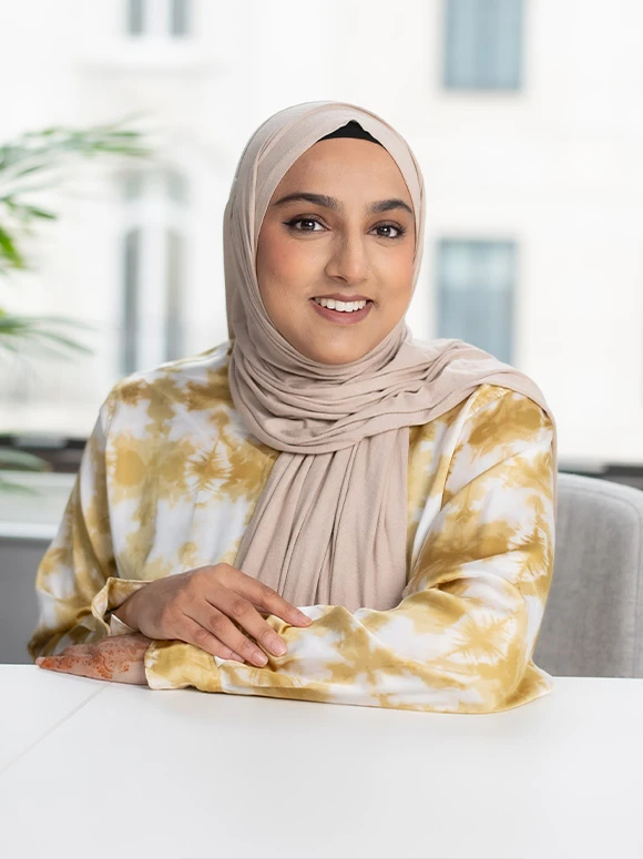 A smiling woman in a yellow and white patterned hijab appears to be sitting at a table, her hands resting on the surface in front of her