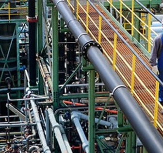 The image shows a large industrial plant with intricate piping and machinery. A worker dressed in blue overalls can be seen walking on a catwalk amidst the equipment