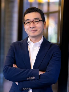 A middle-aged Asian man wearing glasses and a dark suit is standing with his arms crossed, looking at the camera with a serious expression