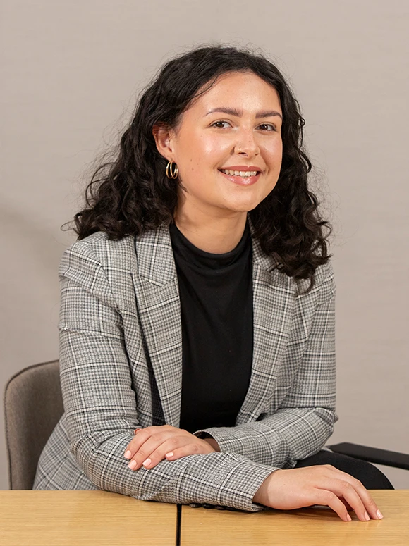 The image shows a smiling woman with dark curly hair wearing a gray and white plaid jacket, seated at a desk and looking directly at the camera