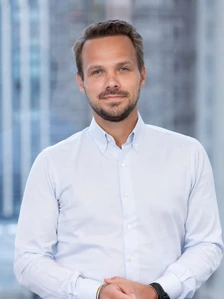 The image shows a smiling man in a white shirt and dark trousers, with a beard and short dark hair, standing in front of a blurred background