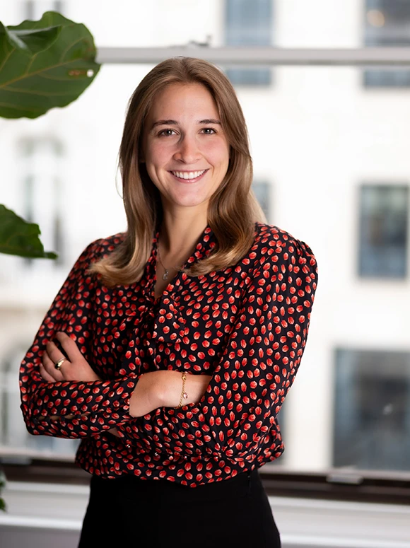 The image shows a smiling woman with long brown hair wearing a red and black patterned blouse, standing in an indoor setting with a potted plant visible