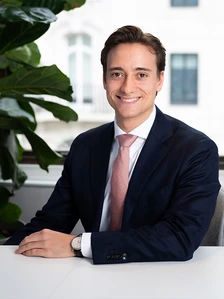 The image shows Konstantin Stoll, a young businessman, sitting at a desk in a professional office setting, smiling confidently while wearing a dark suit and a pink tie