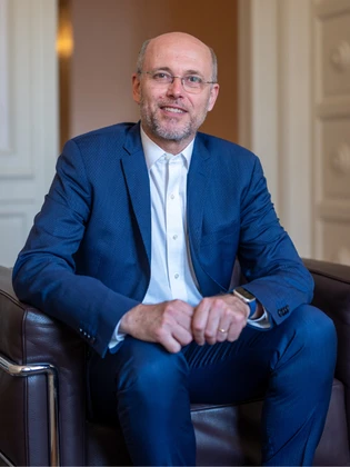 A smiling man with gray hair and a beard, wearing a blue suit and glasses, sitting in a chair and looking directly at the camera