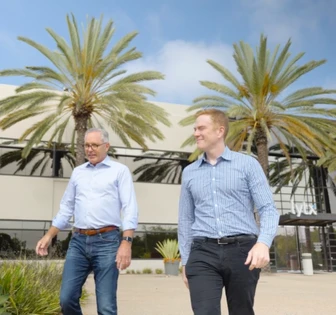 The image shows two men walking together in a sunny outdoor setting with palm trees in the background, suggesting a business partnership or collaboration