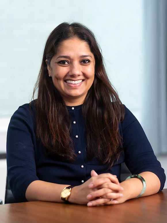 A smiling woman with long dark hair, wearing a navy blue blouse, appears to be Ilina Gunzinger, posing for a professional headshot photograph