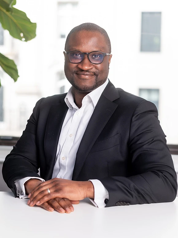 Ansumana Bai Marrow, a confident and professional-looking man in a black suit, is sitting at a desk and smiling directly at the camera