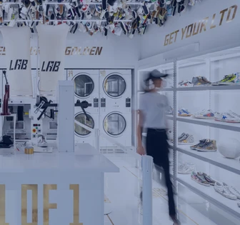 The image shows a well-lit retail space with various shoe and clothing displays, along with a set of washing machines and dryers in the center. A person is visible walking through the space, examining the merchandise