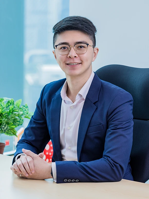 Joshua Sim, a young professional in a blue suit and glasses, is seated at a desk in an office setting, smiling confidently