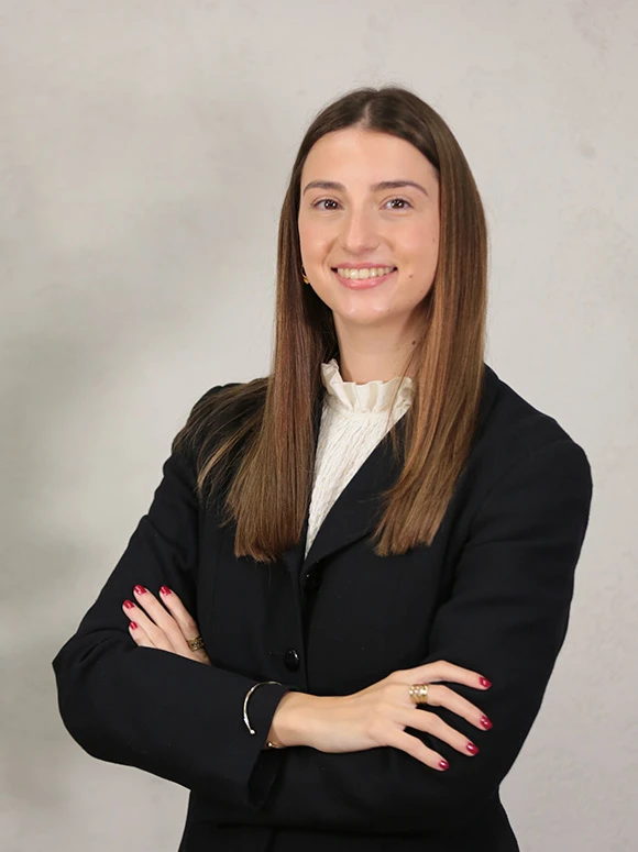 Paula Romero, a smiling young woman with long brown hair, posing in a professional business attire against a light gray background