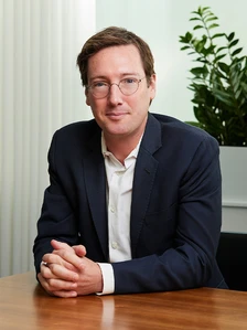 The image shows a middle-aged man wearing a suit and glasses, sitting at a desk and looking directly at the camera. The man appears to be David Kovara, based on the filename