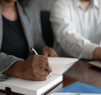 A person, who appears to be working, is writing in a notebook on a desk with other documents and materials present