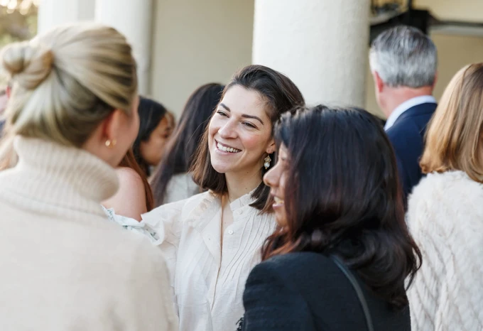 A group of women in formal attire, one smiling brightly in the center, conversing and interacting with each other during what appears to be a social or professional event
