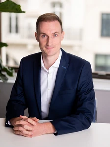 The image shows a smiling, professional-looking man in a navy blue suit, sitting at a desk with a potted plant in the background