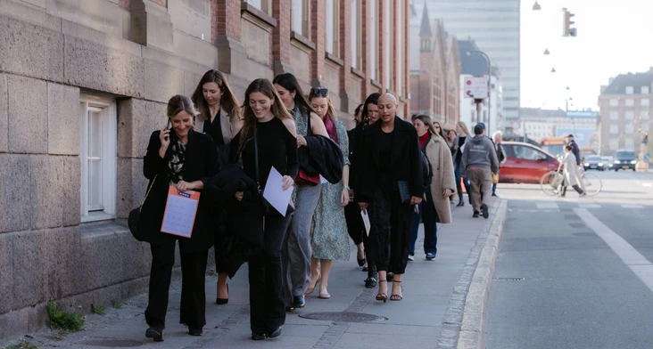 A group of well-dressed individuals walking together on a city street, with one person holding a clipboard and the others appearing to be in conversation