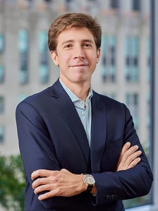 The image shows a young man in a navy blue suit, standing with his arms crossed in front of a blurred building background, with a friendly expression on his face