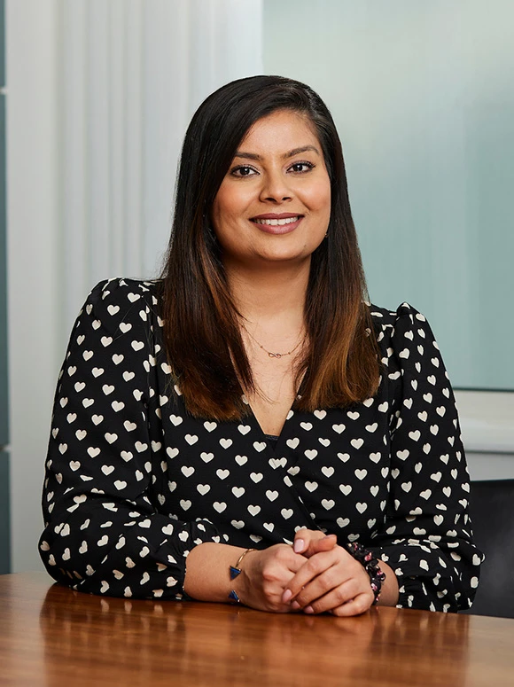 The image shows Priyanka Patel, a smiling woman with dark hair, wearing a black dress with white heart patterns, seated at a desk