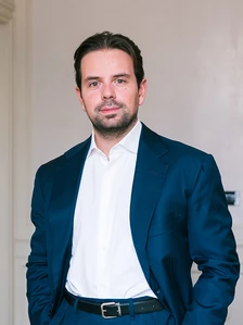 This image shows a well-dressed, middle-aged man with dark hair and a beard, wearing a navy blue suit and white shirt, standing in a professional setting