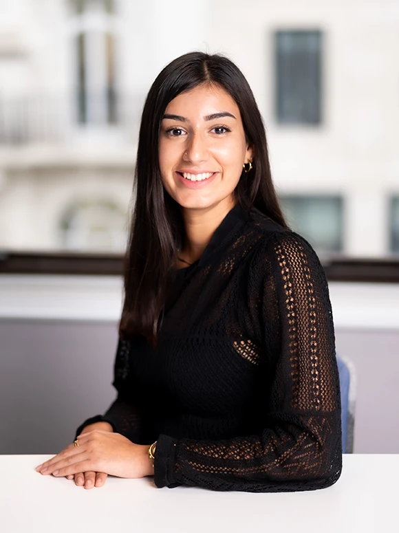 The image shows a young woman with long dark hair, wearing a black lace top, smiling warmly and posing for a portrait in front of a blurred backdrop