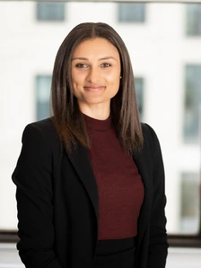 The image shows Courteney Gibson, a woman with long dark hair wearing a burgundy sweater and a black jacket, posing for a professional headshot against a blurred background