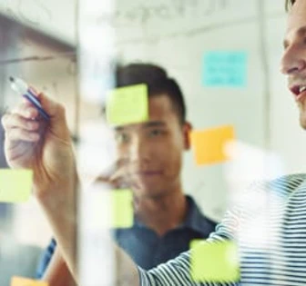 The image shows a group of men working together on a project, writing notes and ideas on a glass board. They appear to be collaborating on a task in a professional setting