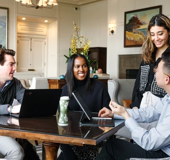 A group of four people, including two women and two men, are having a discussion around a table with laptops and a coffee mug, in a cozy and well-appointed room