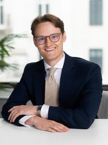 The image shows a smiling young professional man wearing glasses, a suit, and a tie, posing in an office setting. The image is named "Alexander Angelin Web"