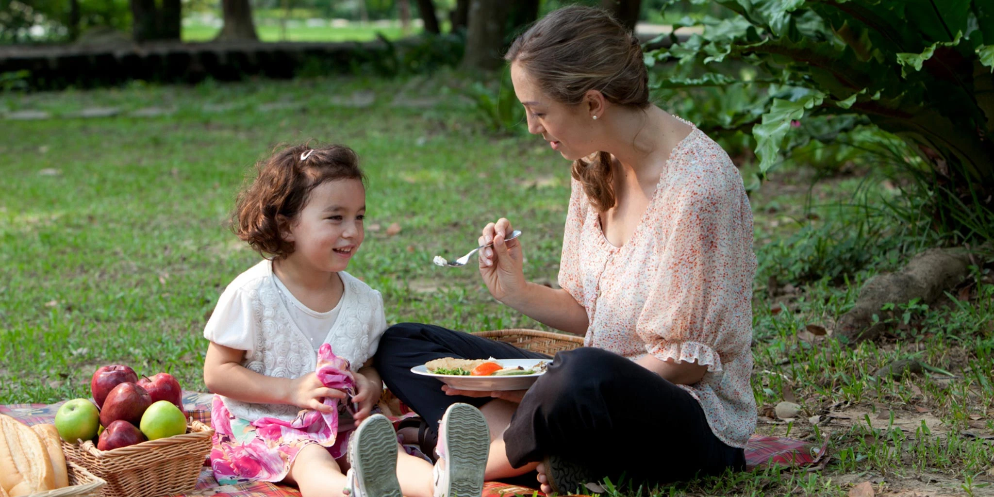 A young woman and a young girl enjoying a picnic in a lush, green garden setting. They appear to be sharing a meal and engaging in a pleasant conversation