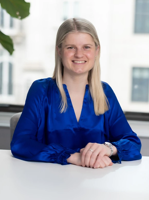 Flora Insley, a young professional woman with long blonde hair, smiling and wearing a blue blouse while seated at a desk