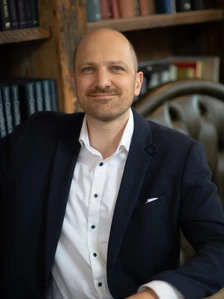 The image shows a headshot of a middle-aged Caucasian man with a bald head and a friendly smile, wearing a navy blue suit and white shirt, and standing in front of a bookshelf