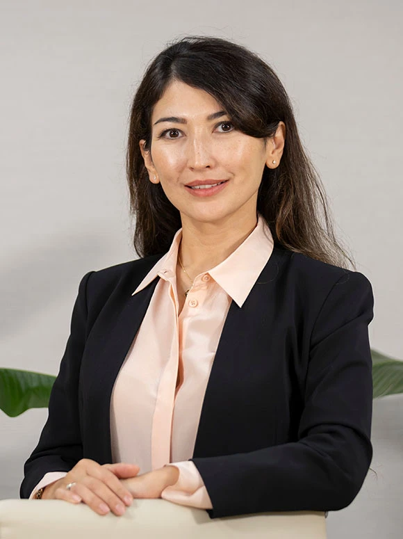 The image shows Indira Ismagulova, a professional woman wearing a black suit jacket and a light peach-colored blouse, sitting at a table and smiling at the camera