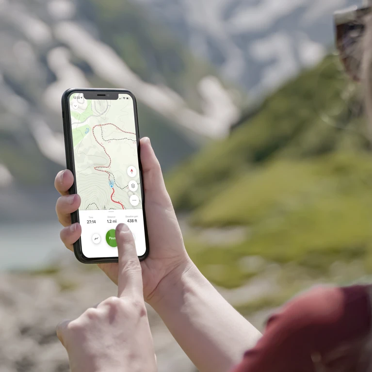 A person's hand holding a smartphone displaying a hiking route map in a scenic mountainous landscape