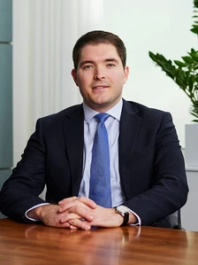 The image shows a well-dressed middle-aged man in a suit and tie, sitting at a desk with his hands folded, in what appears to be a professional office setting
