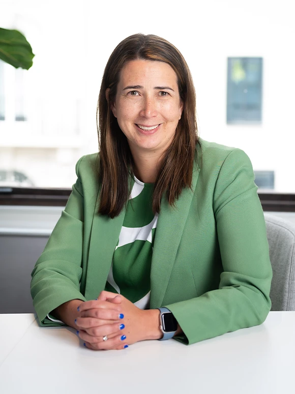 The image shows Annah Currie, a smiling woman in a green blazer, sitting in a professional office setting
