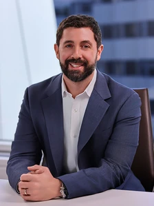 The image shows a smiling man with a beard, wearing a dark blue suit and a white shirt, sitting at a desk and looking at the camera