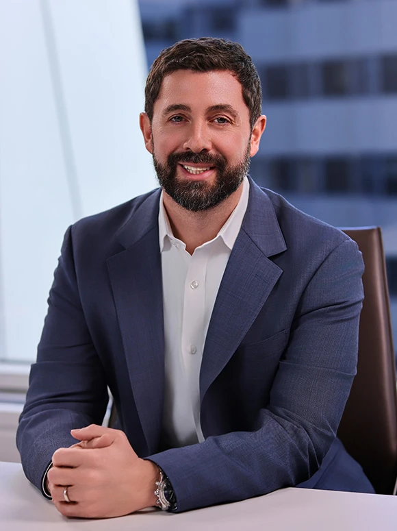 The image shows a smiling man with a beard, wearing a dark blue suit and a white shirt, sitting at a desk and looking at the camera