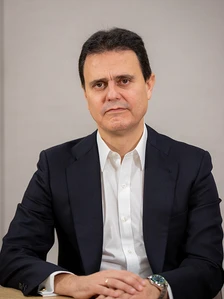 The image shows a middle-aged man with dark hair wearing a dark suit and white shirt, sitting at a table and looking directly at the camera with a serious expression
