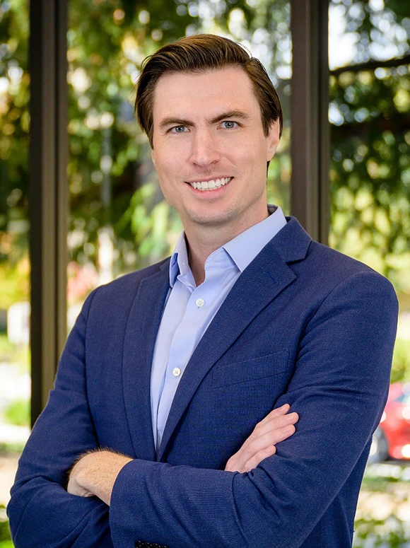 The image shows a headshot of a middle-aged man with short brown hair wearing a blue suit and collared shirt. He is smiling and standing against a blurred background of trees and greenery
