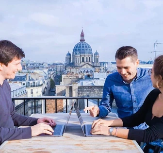 Three people working on laptops on a balcony with a view of the domed Sacré-Cœur Basilica in Paris in the background