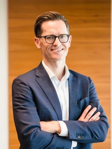Alistair Boyle, a smartly dressed professional, stands confidently with his arms crossed, smiling warmly at the camera in a modern, wooden-paneled setting