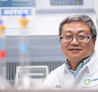 A middle-aged Asian man wearing a white lab coat and glasses, standing in a laboratory with scientific equipment in the background