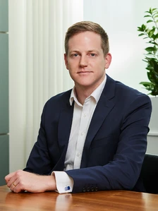 The image shows a well-dressed man with short brown hair sitting at a desk in an office-like setting, looking directly at the camera with a serious expression