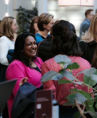 The image shows a group of people, including a smiling woman in a pink jacket, engaged in a conversation in what appears to be a professional or social gathering