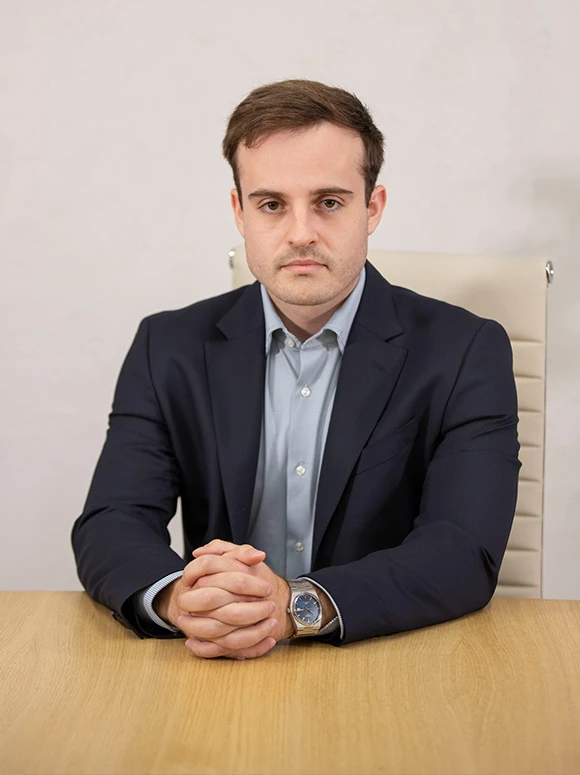 The image shows Alvaro Rojo, a serious-looking man in a navy blue suit, sitting at a desk and looking directly at the camera