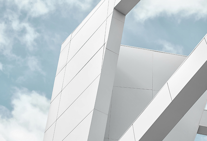 The image depicts a modern, angular building with a striking architectural design featuring intersecting white and gray panels against a cloudy sky