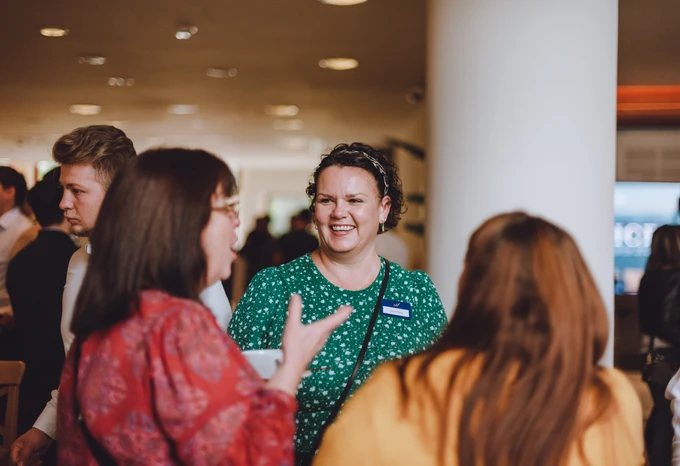 A group of people engaging in conversation at what appears to be a social event, with a smiling woman in a green and white floral dress interacting with two other women