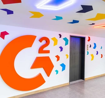 This image shows a colorful, modern hallway with large orange letters "C2G" displayed on the wall, surrounded by various geometric shapes in vibrant colors. People can be seen moving through the hallway in the background