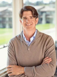 A smiling man with short dark hair wearing a casual beige sweater, photographed in front of a natural landscape