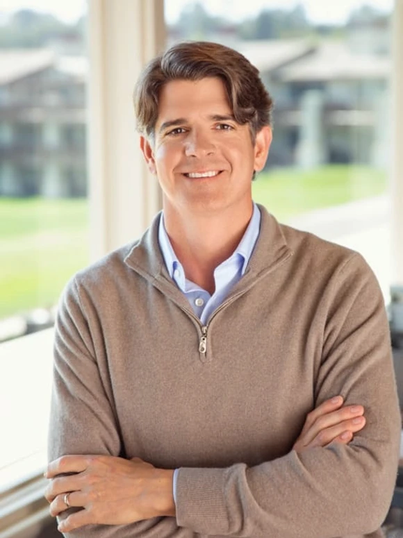A smiling man with short dark hair wearing a casual beige sweater, photographed in front of a natural landscape
